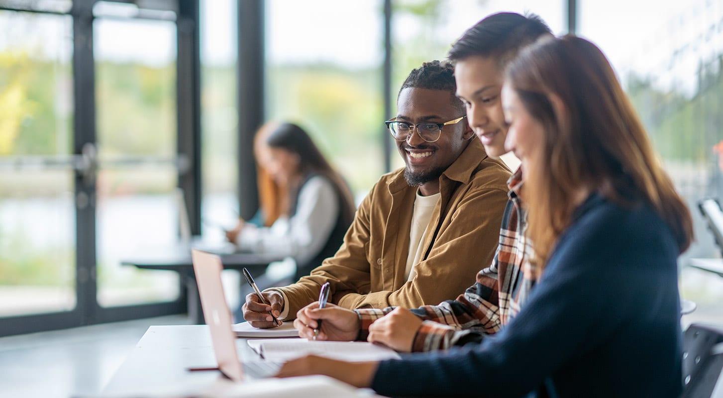 Three students collaborate on a laptop in a classroom, focused on their project and sharing ideas.